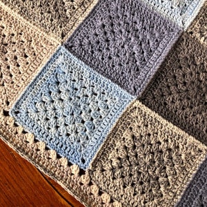 May include: A crocheted blanket with a patchwork design featuring squares in shades of blue, gray, and beige.