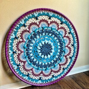Op de afbeelding: Een grote, ronde, gehaakte wanddecoratie met een paarse rand. Het ontwerp heeft een mandala-patroon in tinten blauw, wit en roze.