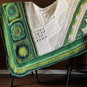 May include: A white crocheted blanket with green and white squares and a green border. The blanket is draped over a black metal chair.