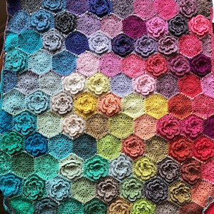 Könnte beinhalten: Eine bunte gehäkelte Decke mit einem sechseckigen Muster und blütenförmigen Motiven. Die Decke ist in verschiedenen Farben gehalten, darunter Rosa, Lila, Blau, Grün, Gelb und Orange.
