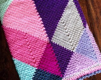 May include: A colorful crocheted blanket with a geometric pattern in shades of pink, purple, blue, and green. The blanket has a scalloped edge.
