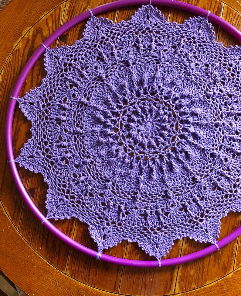 Op de afbeelding: Een paarse gehaakte doily is gespannen over een paarse plastic hoepel.