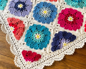 May include: A crocheted blanket with a white border and colorful squares featuring flower designs. The squares are in shades of blue, pink, red, and green.