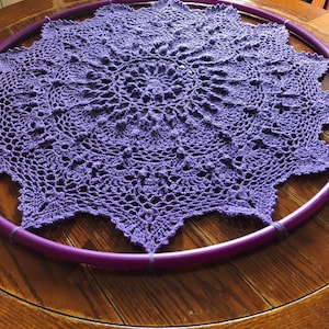 Op de afbeelding: Een paarse gehaakte doily is gespannen over een paarse hoepel, rustend op een houten tafel.