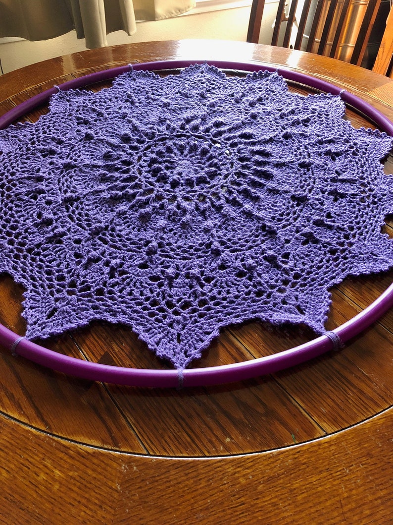 Op de afbeelding: Een paarse gehaakte doily is gespannen over een paarse hoepel, rustend op een houten tafel.