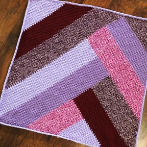 Pode incluir: Uma manta de crochê com um padrão chevron em tons de roxo, rosa e bordô.