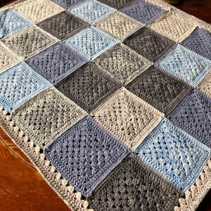 May include: A crocheted baby blanket with a patchwork design in shades of blue, gray, and beige. The blanket has a crocheted border.