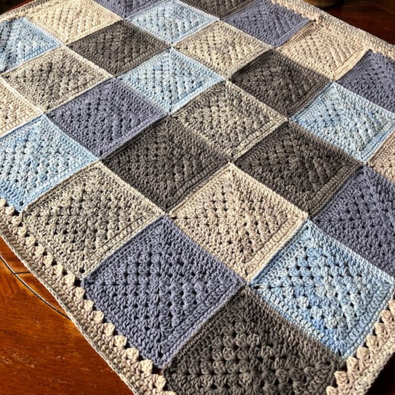 May include: A crocheted baby blanket with a patchwork design in shades of blue, gray, and beige. The blanket has a crocheted border.