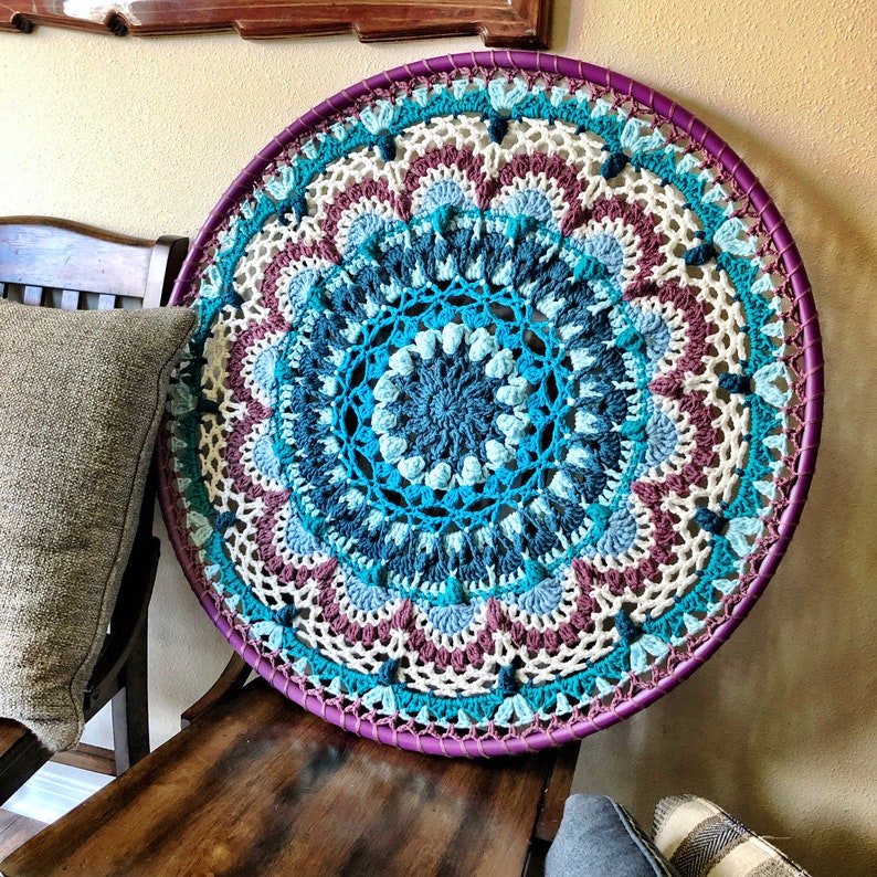 Op de afbeelding: Een grote, ronde, gehaakte wandhanger met een mandala-ontwerp in tinten blauw, turquoise, wit en paars. De mandala is omgeven door een witte gehaakte rand en een paarse houten ring.