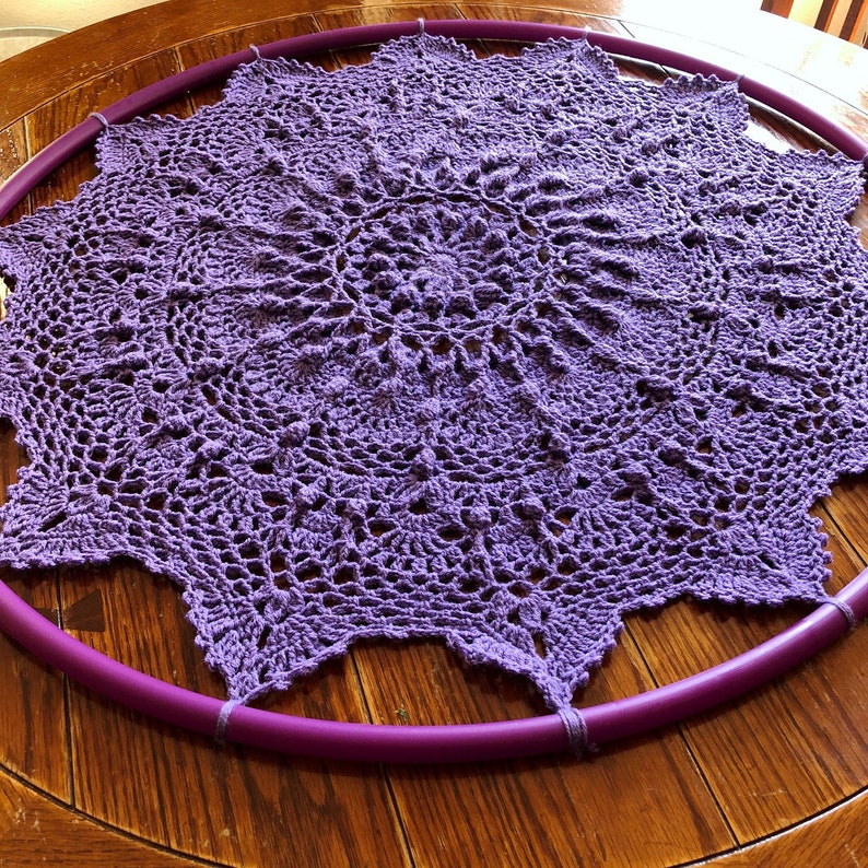 Op de afbeelding: Een grote, paarse gehaakte doily is gespannen over een paarse hoelahoep. De doily heeft een delicaat, ingewikkeld ontwerp.