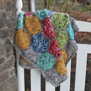May include: A colorful crocheted blanket with a hexagonal pattern in shades of red, green, blue, yellow, and gray. The blanket is draped over a white picket fence.