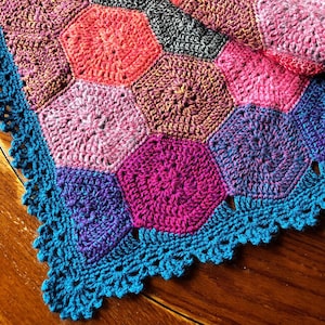 Puede incluir: Una manta de ganchillo con un borde turquesa y un patrón hexagonal en tonos de rosa, morado y rojo.
