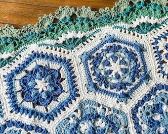 May include: A crocheted blanket with a scalloped edge in shades of blue and green. The blanket is made up of repeating hexagonal patterns.