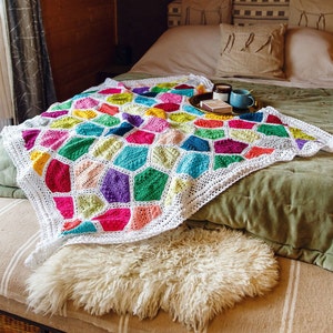 May include: A white crocheted blanket with a colorful geometric pattern in shades of pink, green, blue, yellow, and orange. The blanket is draped over a bed with a white pillow and a brown throw pillow.