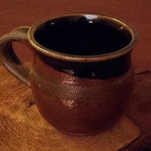 May include: A brown ceramic mug with a dark brown rim and a handle. The mug is sitting on a wooden surface.