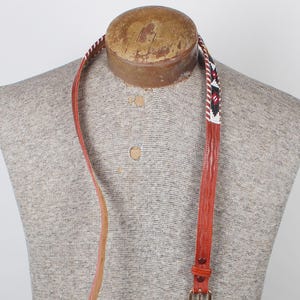 May include: A thin orange leather belt featuring a section of intricate white, black, and red geometric beadwork. The belt includes a simple metal buckle and a matching leather keeper loop, displayed on a textured, neutral-toned mannequin torso.