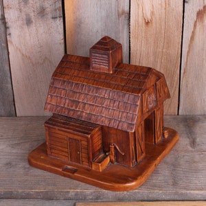 May include: A wooden barn shaped trinket box with a hinged door and a small cupola on the roof. The box is made of dark brown wood and has a rustic appearance.