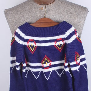 May include: A blue knitted sweater with a white and red geometric pattern. The sweater has a crew neck and long sleeves.
