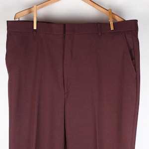 May include: Burgundy-colored dress pants hanging on a wooden hanger. The pants have a classic, straight-leg design with front pockets and a button closure. The fabric appears to be a textured weave.