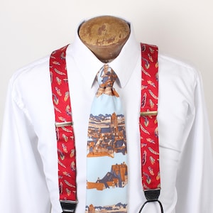 May include: A set of red suspenders featuring a multi-colored feather pattern with silver-toned metal adjusters and black leather attachments, paired with a light blue necktie displaying a repeating landscape print of buildings and a river.