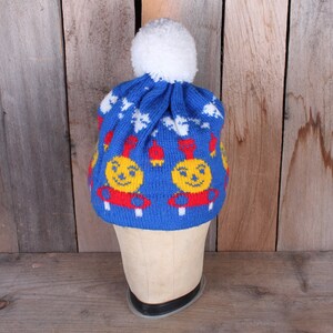 May include: Blue knitted hat with a white pom pom and a repeating pattern of red, yellow, and white smiling faces.