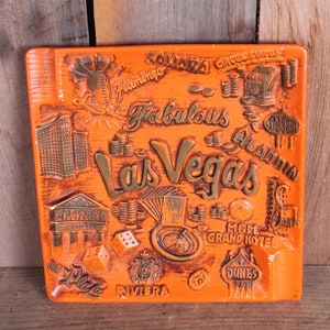 May include: Orange ceramic ashtray with a raised design of various Las Vegas landmarks and casinos, including the Flamingo, Aladdin, and the MGM Grand Hotel. The text "Fabulous Las Vegas" is in gold lettering.