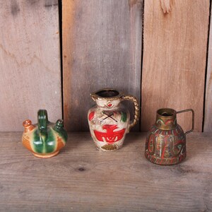 May include: Three small decorative ceramic and metal pitchers with painted designs. The pitchers are green, white, and gold with red and black accents. The pitchers are arranged on a wooden surface.