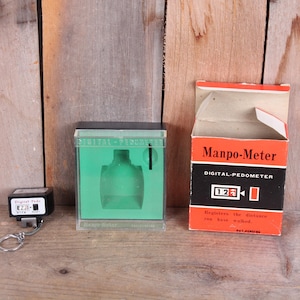 May include: A vintage digital pedometer, a clear plastic case with a green interior, and an open box labeled "Manpo-Meter" are displayed on a wooden surface. The pedometer is black with a silver key ring.