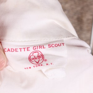Puede incluir: Camisa blanca de uniforme de Girl Scout Cadete con una etiqueta roja que dice "Cadette Girl Scout" y "adette" con un logotipo de tr&eacute;bol. La etiqueta tambi&eacute;n dice "New York, N.Y."