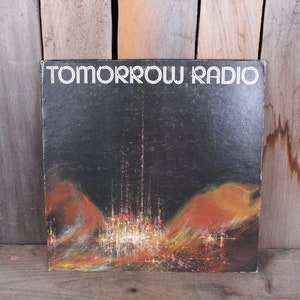 May include: A black vinyl record album cover with the title "TOMORROW RADIO" in white text. The cover art features a stylized cityscape with a tall tower in the center, surrounded by orange and yellow hues.