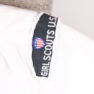 Puede incluir: Camiseta blanca de uniforme de Girl Scouts USA con un parche blanco y negro con el texto "Girl Scouts USA" y un escudo rojo, blanco y azul con estrellas.