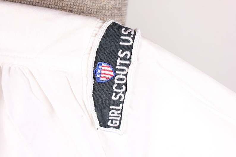 Puede incluir: Camiseta blanca de uniforme de Girl Scouts USA con un parche blanco y negro con el texto "Girl Scouts USA" y un escudo rojo, blanco y azul con estrellas.