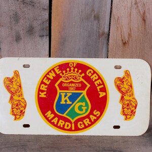 May include: White metal license plate with a red, yellow, and blue circular emblem that reads "Krewe of Grela Mardi Gras" and "Organized 1947" with a crown and "KG" inside the emblem. Two yellow figures with red faces are on either side of the emblem.