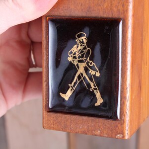 May include: Wooden box with a black and gold emblem. The emblem features a gold figure of a man in a top hat and coat, walking with a cane. The box is held by a hand.