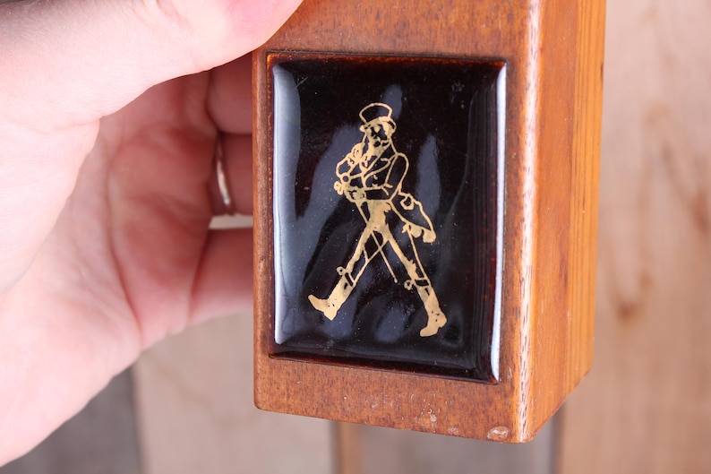 May include: Wooden box with a black and gold emblem. The emblem features a gold figure of a man in a top hat and coat, walking with a cane. The box is held by a hand.