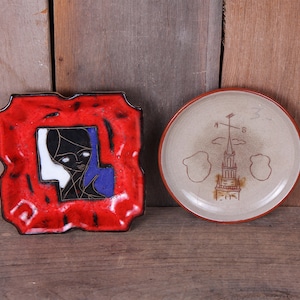 May include: Two ceramic plates, one red with a black and white portrait of a woman, the other beige with a brown drawing of a building and a compass.