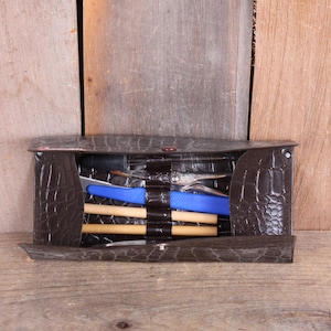 May include: A dark brown, faux-leather tool kit with a crocodile skin texture. The kit is open, revealing various tools, including pliers, a blue tool, and wooden dowels. The background is a wooden surface.