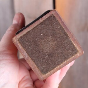 May include: A square, wooden object with a brown felt bottom and a dark edge. The object is held in a hand, with the focus on the felt and the wooden frame. The object appears to be a small box or a base for something.