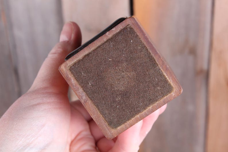 May include: A square, wooden object with a brown felt bottom and a dark edge. The object is held in a hand, with the focus on the felt and the wooden frame. The object appears to be a small box or a base for something.