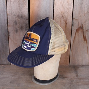 May include: A navy blue and tan trucker hat with a patch that reads "LOWRANCE". The patch features a mountain and fish design. The hat has a mesh back and a curved brim.