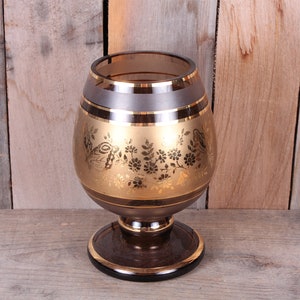 May include: A dark brown glass vase with a gold rim and gold floral pattern. The vase has a wide base and a round body.