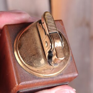 May include: A vintage lighter with a brown wooden base and a gold-colored metal top. The lighter's mechanism is also gold-toned, with a textured striker. The design is rectangular with rounded edges.