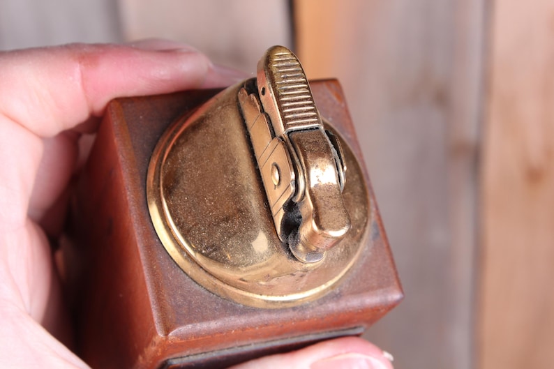 May include: A vintage lighter with a brown wooden base and a gold-colored metal top. The lighter's mechanism is also gold-toned, with a textured striker. The design is rectangular with rounded edges.