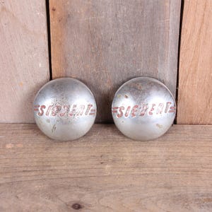 May include: Two silver-colored, domed metal objects with the word "SIEBERT" in red lettering. The objects are slightly weathered and rest on a wooden surface. The items appear to be vintage automotive parts.