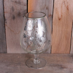 May include: A clear glass vase with a silver floral design. The vase has a round body and a wide base.