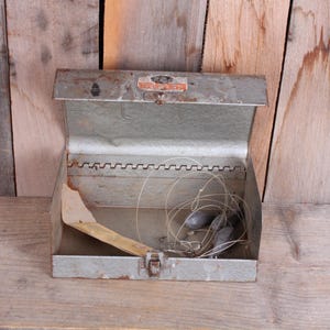 May include: An open, vintage metal tackle box with a hinged lid. Inside are fishing weights, line, and a wooden piece. The box is weathered with a latch closure and sits on a wooden surface.