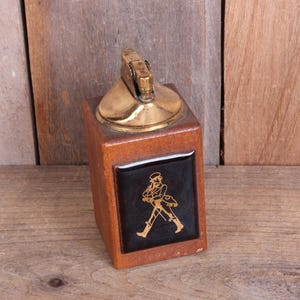 May include: Vintage wooden lighter with a brass top and a black and gold emblem featuring a walking figure. The lighter is rectangular and sits on a wooden surface.