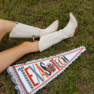 May include: Cream-colored cowboy boots with decorative stitching and a pointed toe rest on the grass near a Texas Tech pennant. The pennant is white with the words "TEXAS TECH" in orange and black.