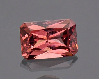 May include: A rectangular, faceted gemstone with a reddish-pink hue. The gemstone is cut in a step-cut style, with many facets that reflect light.