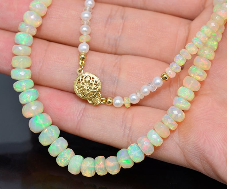 May include: A delicate necklace made with white opal beads and a gold clasp. The necklace is displayed on a hand.
